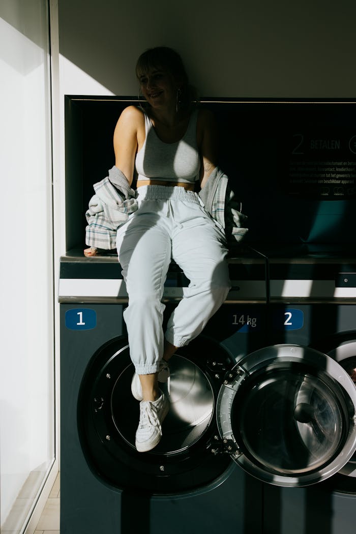 A trendy young woman sitting on a washing machine enjoying sunlight indoors.